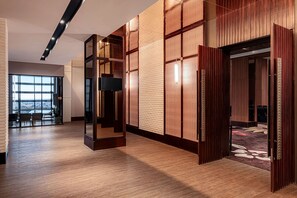 Eine großzügige Hotelhalle mit Holzvertäfelung, einem großen Flachbildfernseher und Blick auf die Stadt durch hohe Fenster.