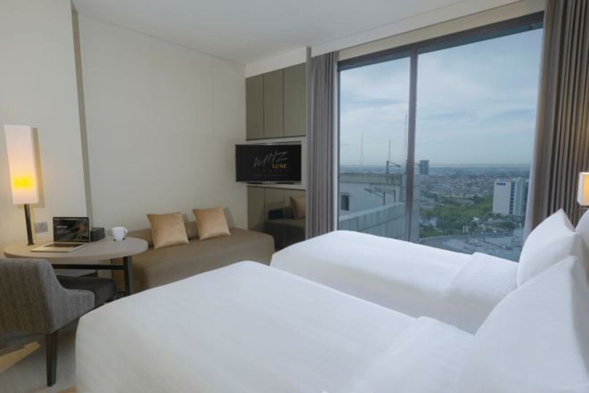 A hotel room with two beds, a TV, a desk with a chair, a sofa, and a large window offering a city view.