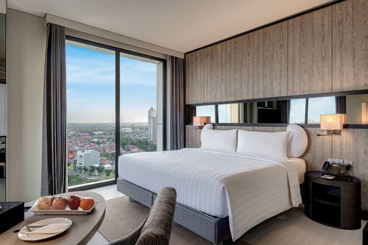 Une chambre d’hôtel moderne dotée d’un grand lit, d’un coin repas avec une table et des chaises, et offrant une vue sur la ville grâce à de grandes fenêtres.