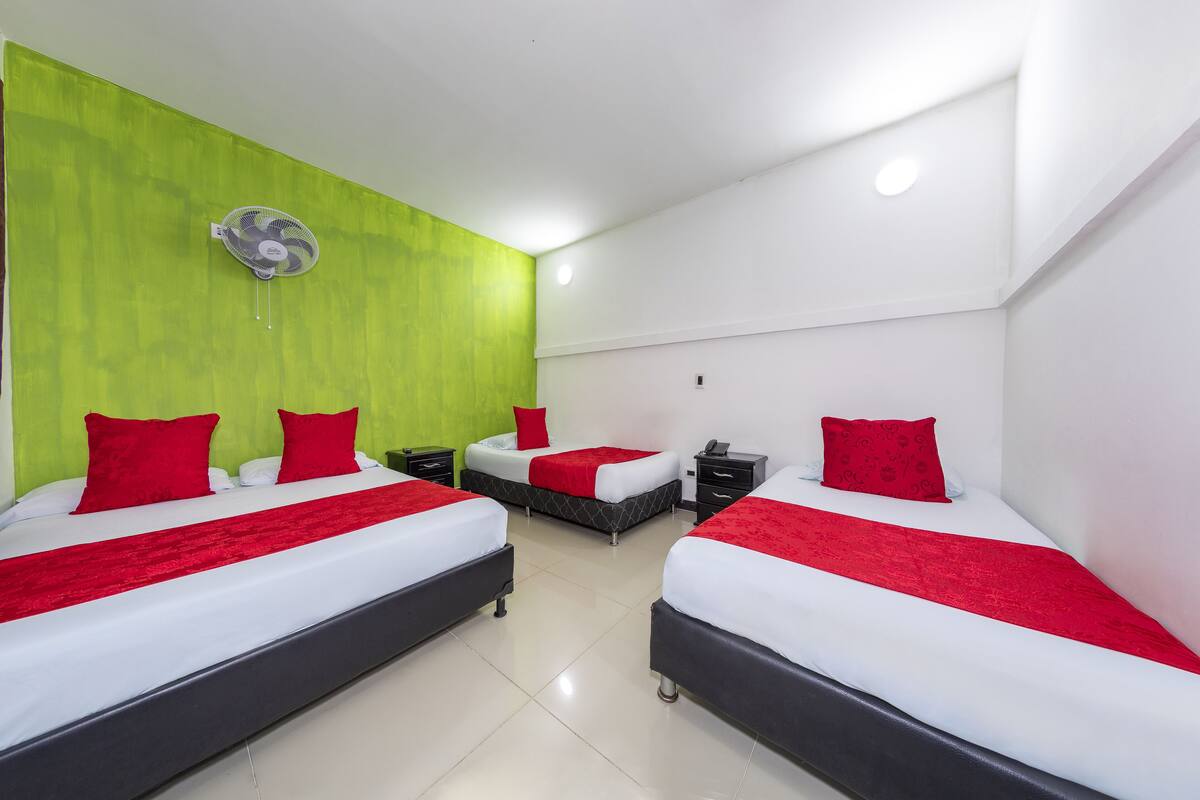 Ein Hotelzimmer mit zwei Betten, jeweils mit roten Kissen und einer roten Bettdecke. An einer Wand ist ein grüner Akzentfarbton zu sehen, und eine Deckenventilator ist angebracht.