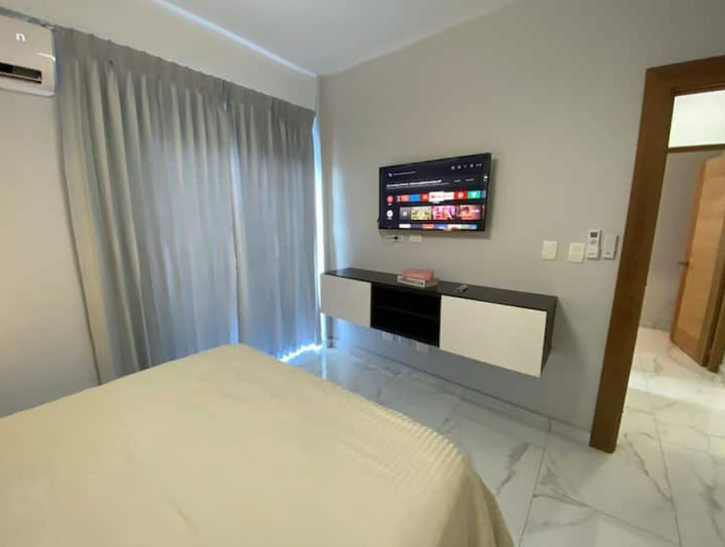Una habitación de hotel con una cama, un televisor montado en la pared, un estante pequeño y una puerta.