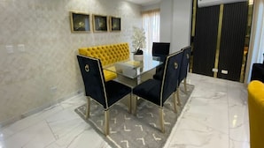 Een eetgedeelte met een glazen tafel, zwarte bekleden stoelen, een geel gestofte bank en een marmeren vloer.
