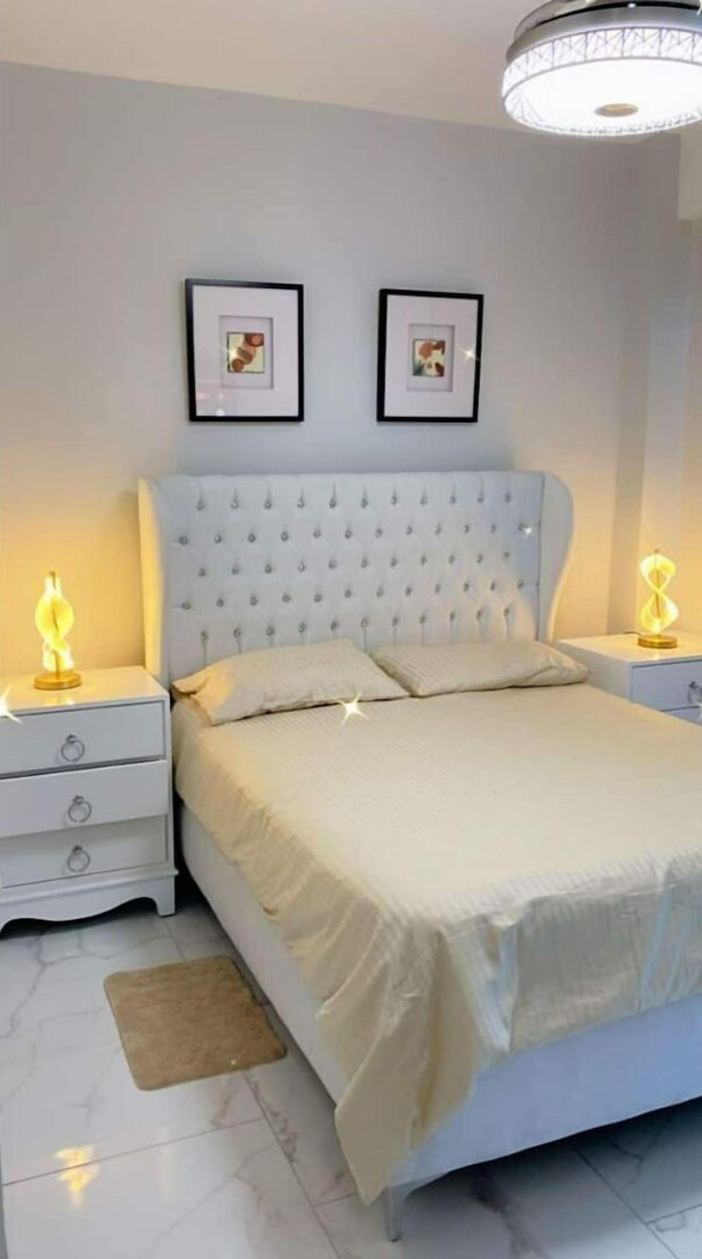Een slaapkamer met een gestoffeerd hoofdeinde, twee nachtkastjes met lampen, een bed met een beige sprei, en twee ingelijste foto’s aan de muur.