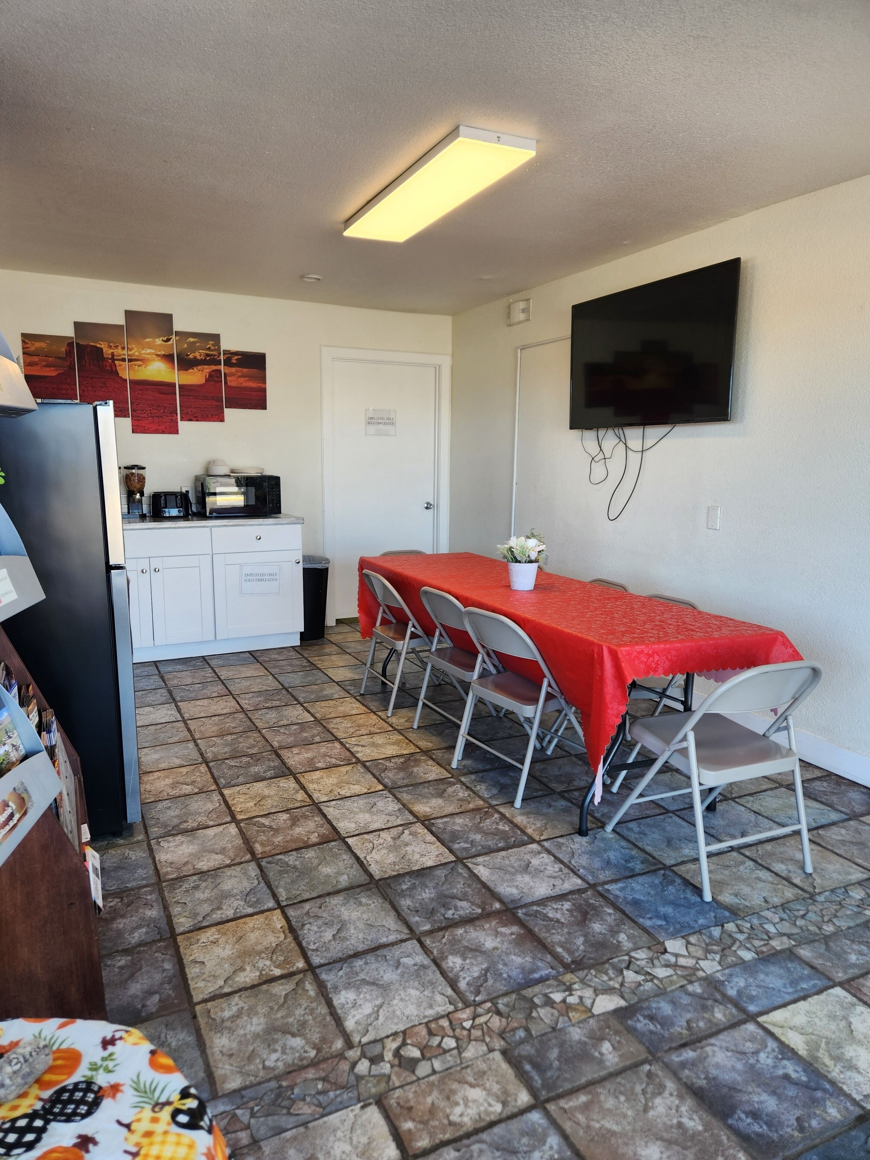 Una cocina compacta con refrigerador, microondas y tostadora, un área de comedor con una mesa y sillas rojas, y un televisor montado en la pared.