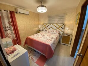 Une chambre à coucher comprenant un lit, des tables de chevet, une commode, un miroir, une fenêtre avec des rideaux et un appareil de climatisation.