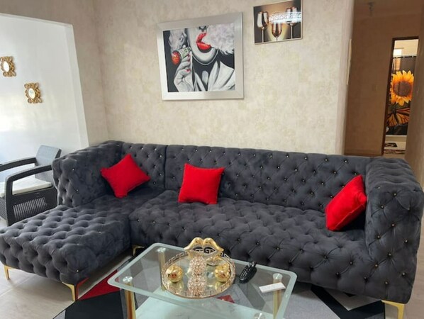 Un salon moderne avec un canapé capitonné gris foncé, des coussins rouges et une table basse en verre agrémentée d’objets décoratifs.
