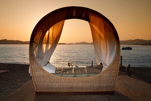 Una cama redonda de mimbre con cortinas ligeras, una mesita con bebidas y vistas al mar y a las montañas al atardecer.