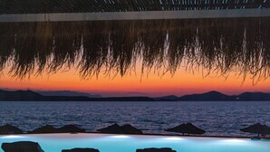 Una piscina infinita con sillas de descanso, vistas al atardecer y sombrillas de paja.