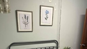 Un lit bien fait, recouvert d’une courtepointe blanche matelassée, avec deux oreillers gris et une tête de lit en métal. Deux reproductions encadrées de motifs botaniques sont accrochées au mur au-dessus du lit.