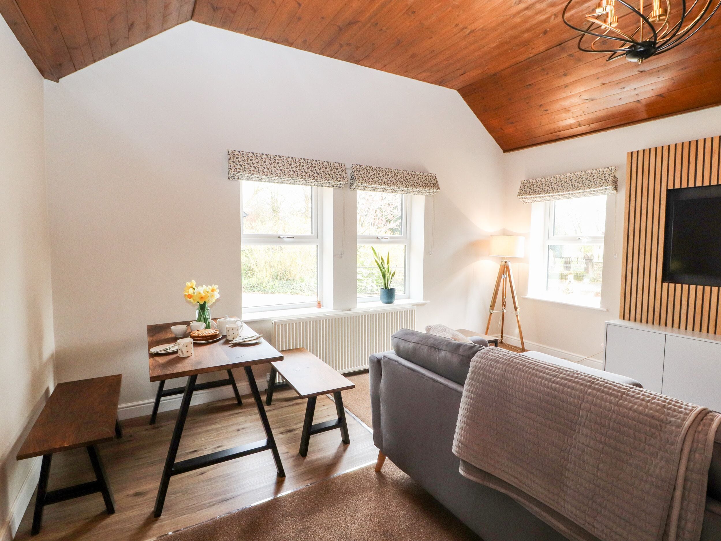 A cozy room with a wooden ceiling, a grey sofa, a small table with a vase of flowers, and a lamp.