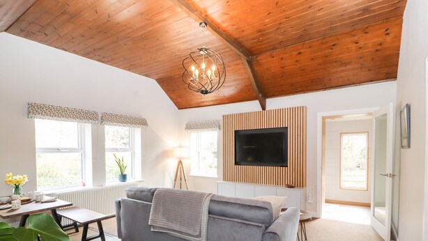 Een gezellige woonkamer met een grijze bank, houten plafond en een flatscreen televisie aan de muur.