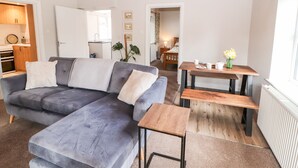Een moderne woonkamer met een grijze modulaire bank, een houten salontafel en een klein bijzettafeltje. Achtergrond toont een keuken met houten kasten en een aanrecht.
