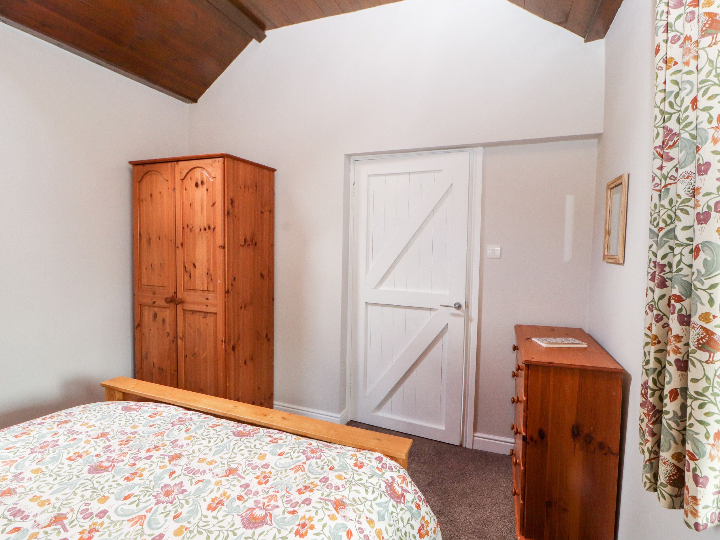 A bedroom with a bed, a wooden wardrobe, a white door, and a small wooden cabinet.