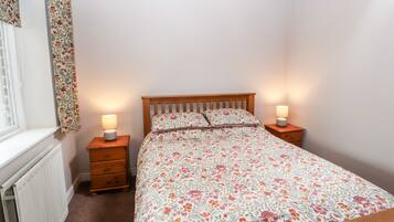 A bedroom with a wooden bed, floral bedding, two bedside lamps, a wooden nightstand, a radiator, and a window with a floral curtain.
