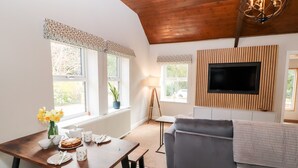 Een moderne woonkamer met een houten plafond, een grijze bank, een houten eettafel met een bloevas, en een flatscreen televisie bevestigd aan een houten wandpaneel.