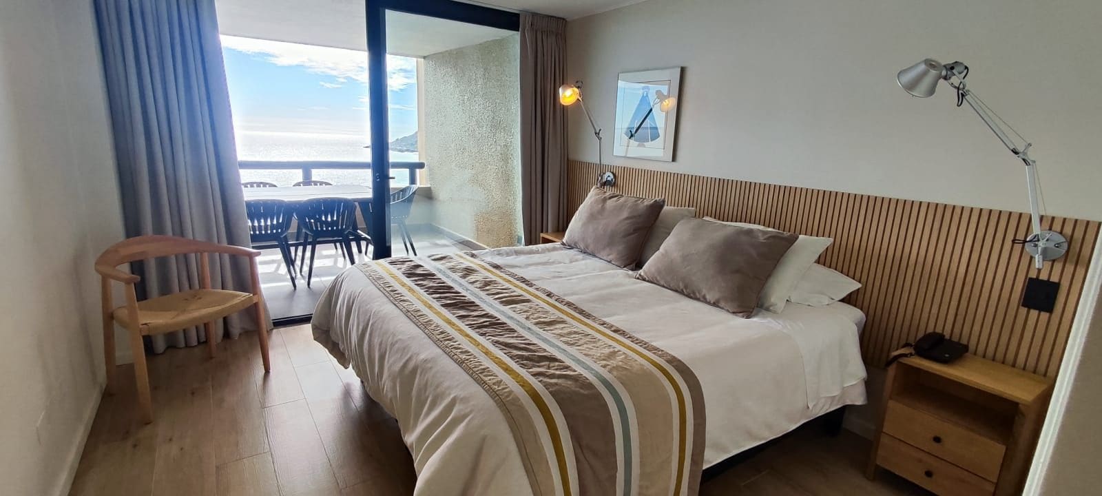 A hotel room with a bed, bedside table, lamp, and a view of the sea through an open door.