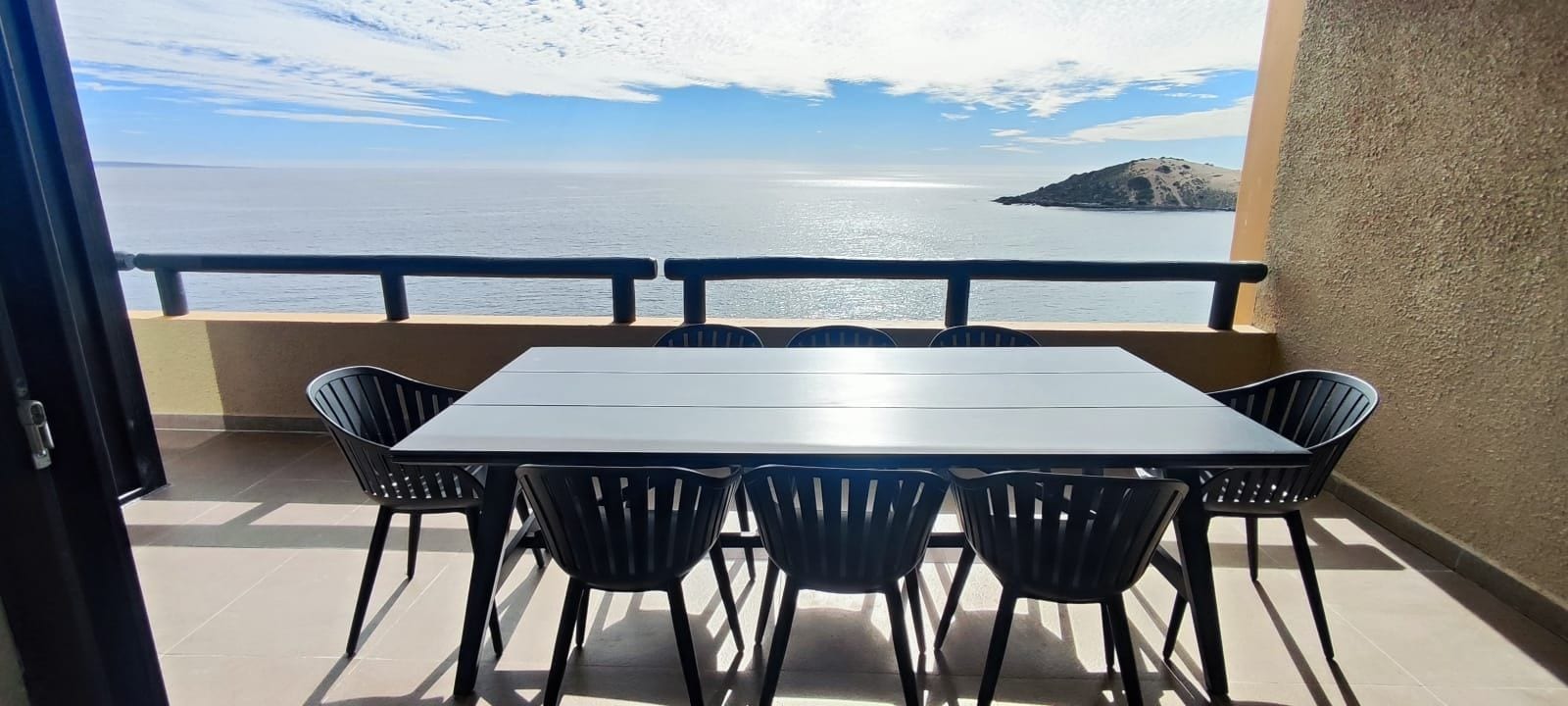 A balcony with a table and chairs, overlooking a body of water and a distant island.