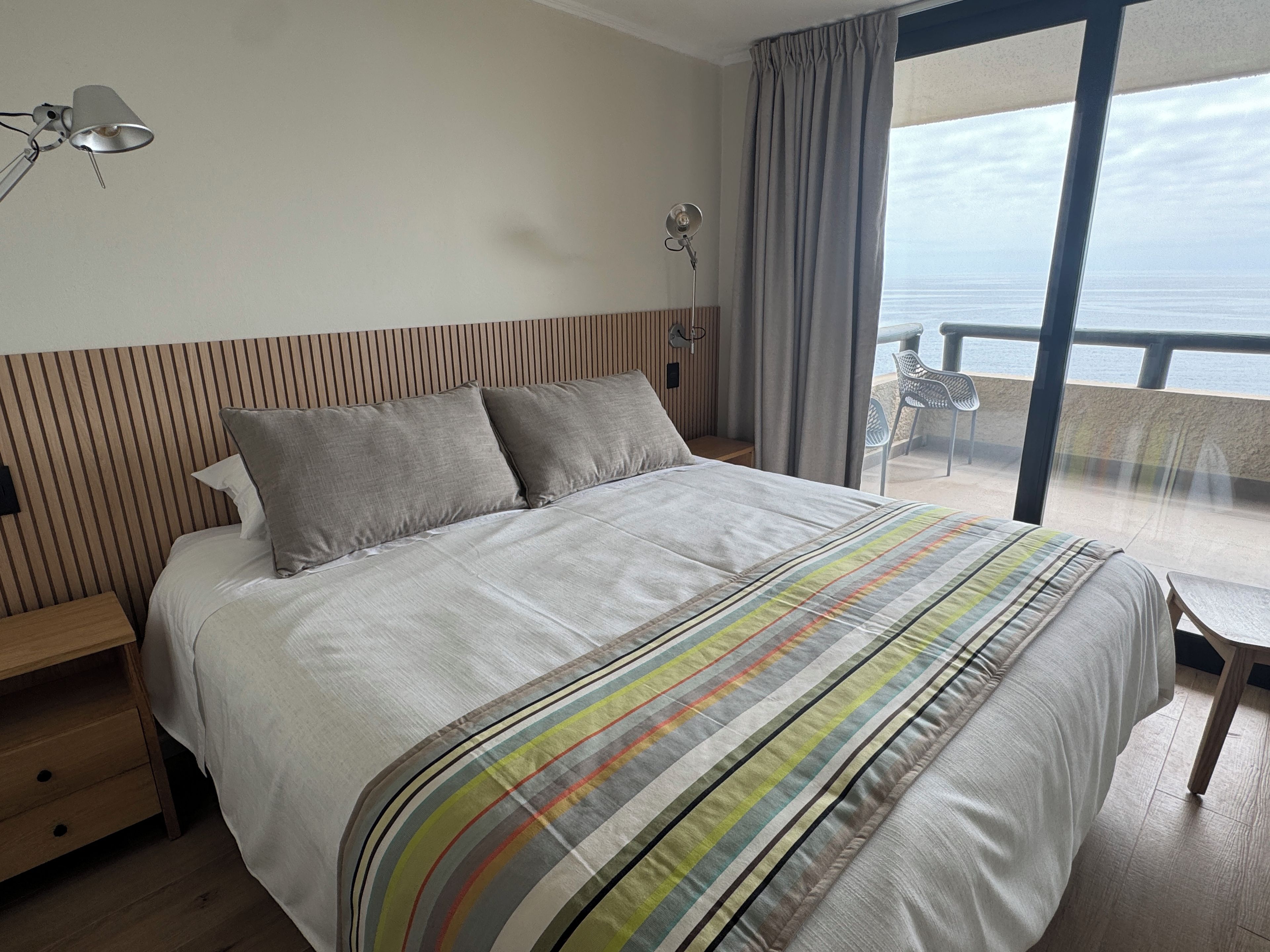 A hotel room with a bed, a bedside table, a lamp, a view of the sea through a balcony.