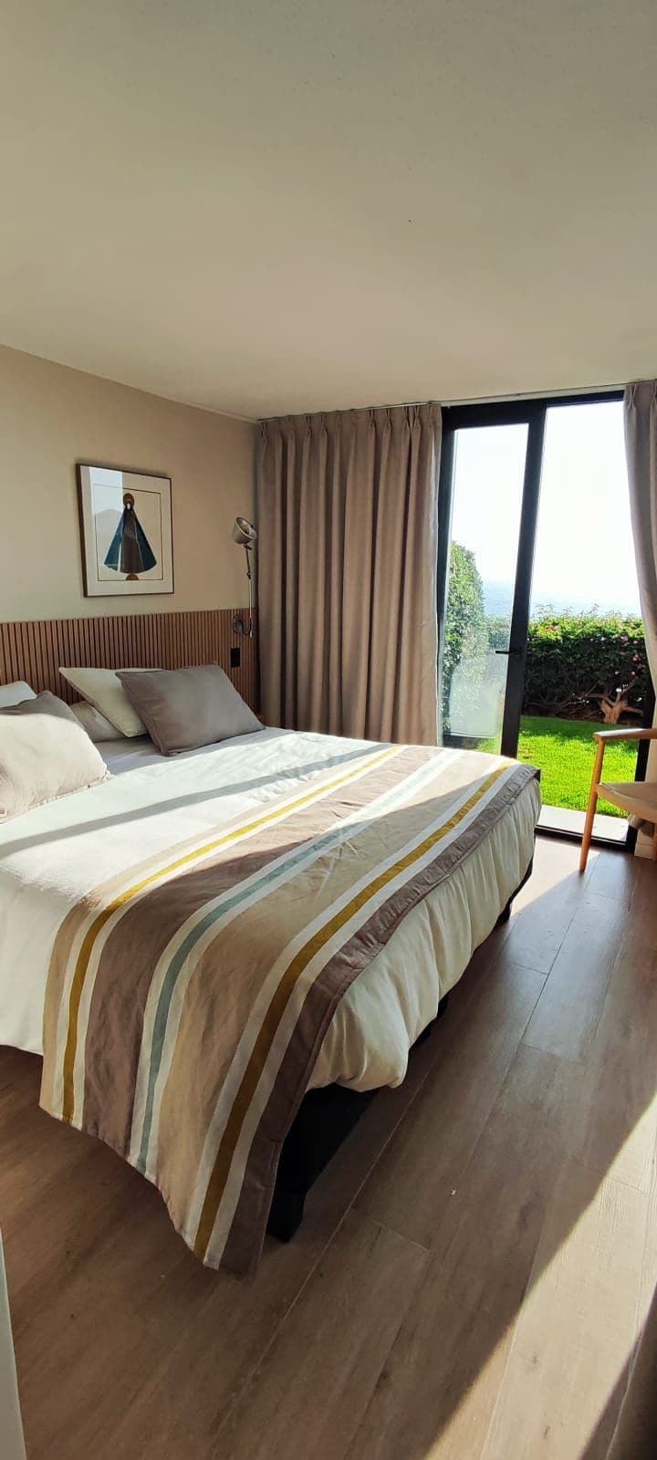 A bedroom with a large bed, a nightstand, a lamp, and a view of a garden through a sliding glass door.