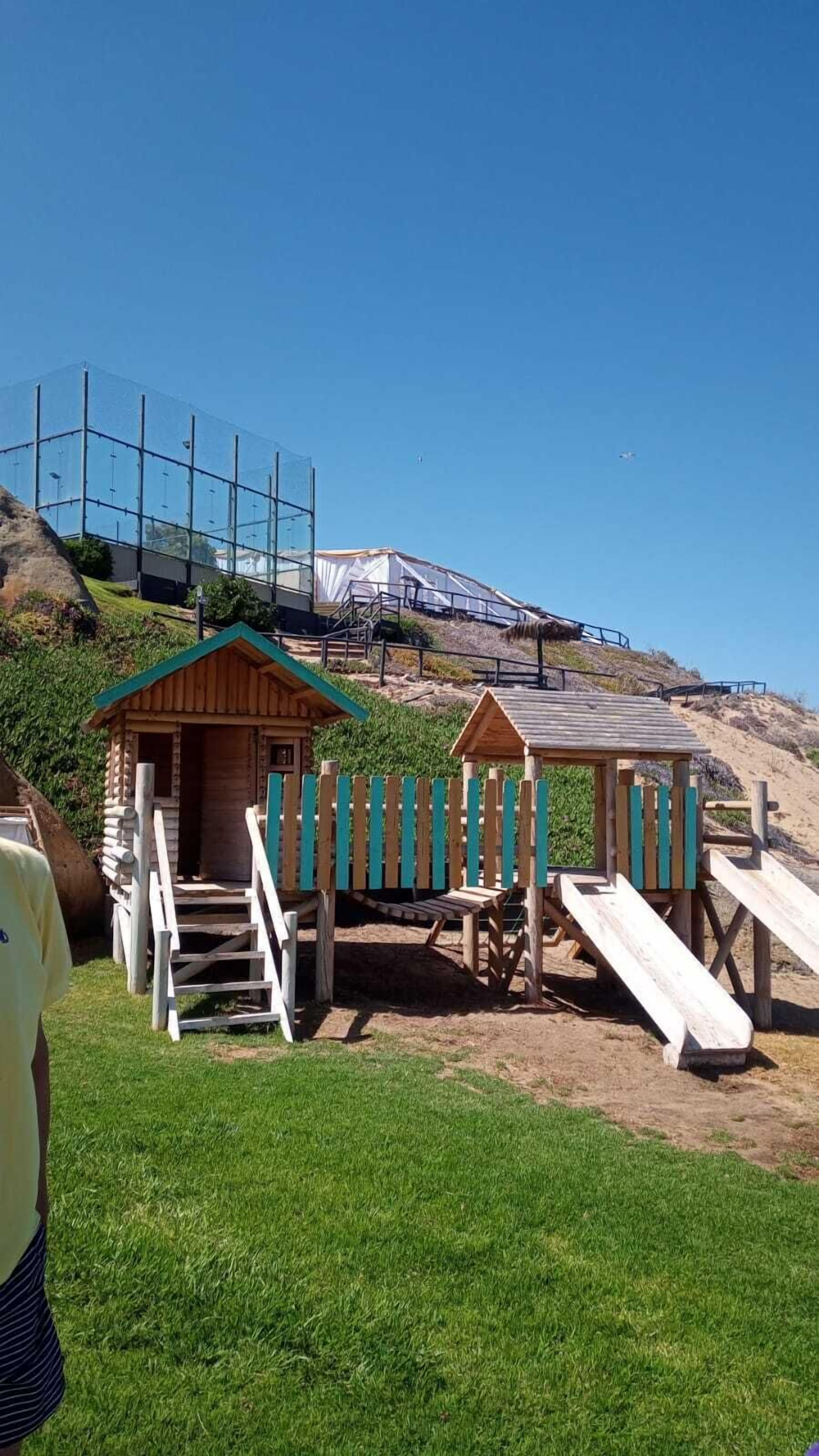 Uma área de praia com casinhas de madeira e um escorrega, cercada por uma cerca e um monte gramado.