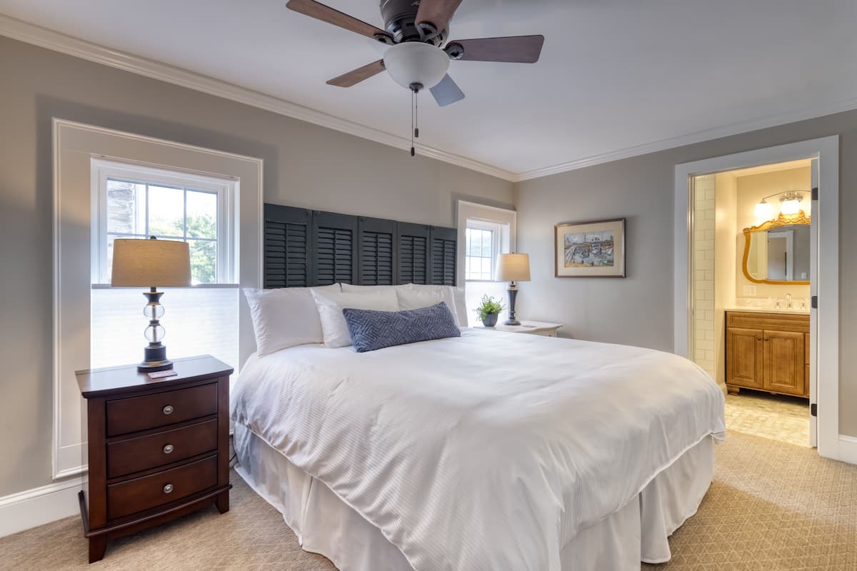 A bedroom with a large bed, a nightstand with drawers, a ceiling fan, and a bathroom visible through an open door.