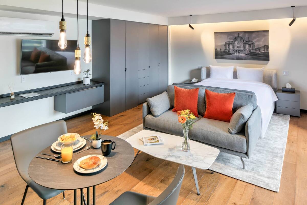 A modern hotel room with a grey sofa, a dining table with breakfast, a TV mounted on the wall, and a large bed with a nightstand.