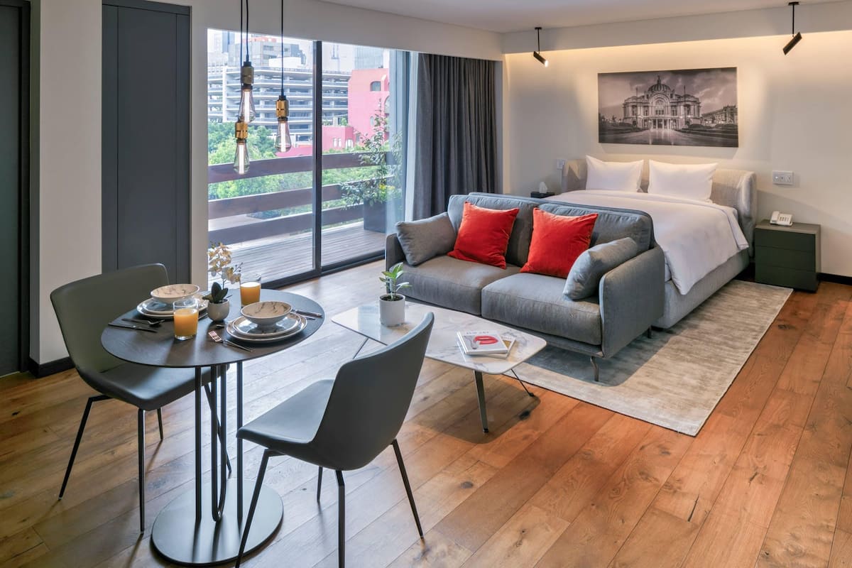 A modern hotel room with a large bed, grey sofa, and a dining area with a table and chairs.