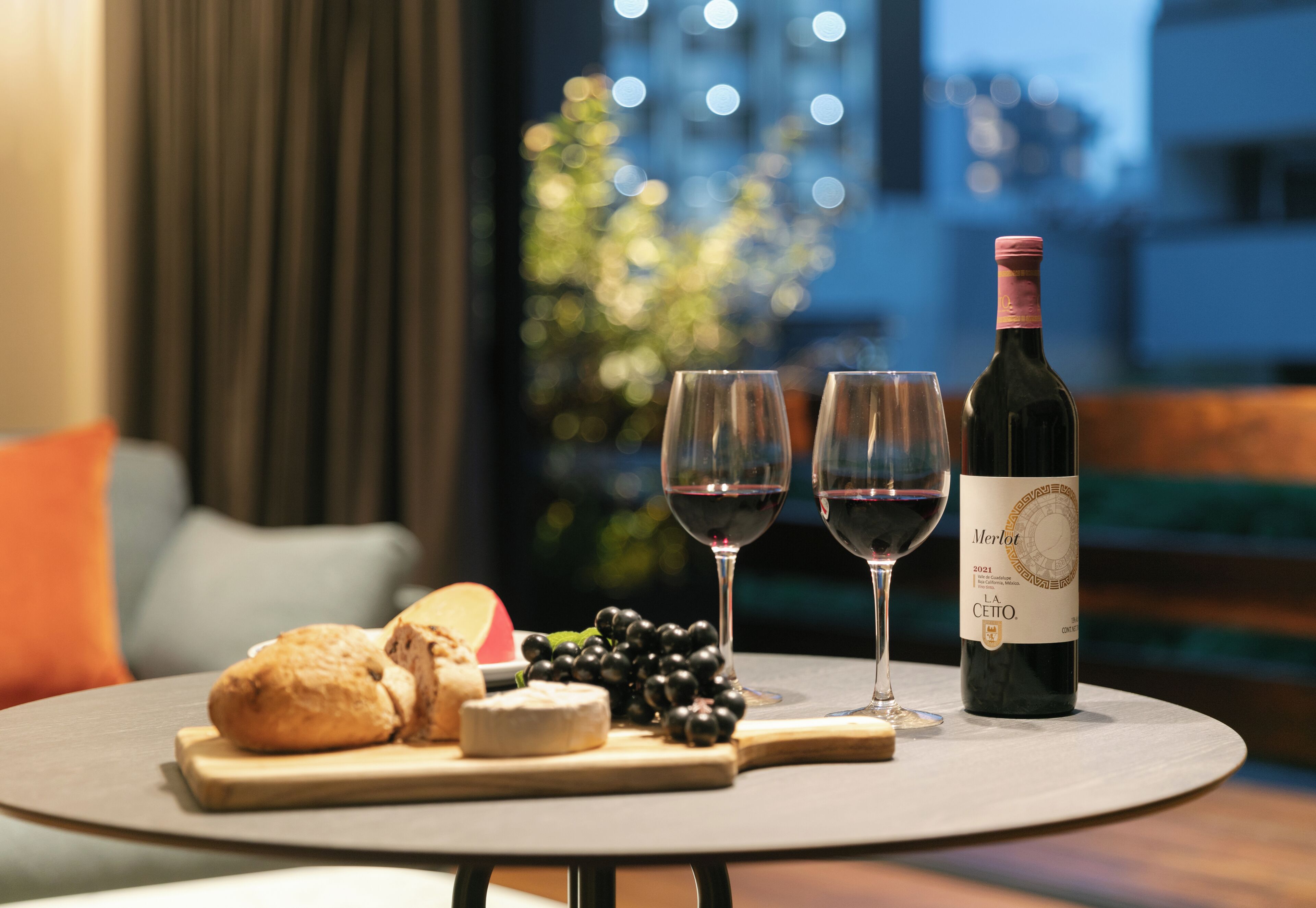 A table with a bottle of Merlot, two glasses of wine, a loaf of bread, a cheese, and grapes.