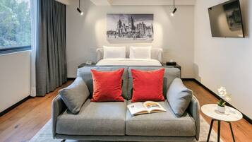 Un dormitorio moderno con un sofá gris, almohadas rojas, una cama con sábanas blancas, un cuadro de un paisaje urbano, una mesita con una planta y un amplio ventanal con cortinas.