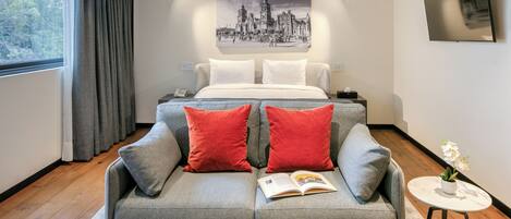 Un dormitorio moderno con un sofá gris, almohadas rojas, una cama con sábanas blancas, un cuadro de un paisaje urbano, una mesita con una planta y un amplio ventanal con cortinas.