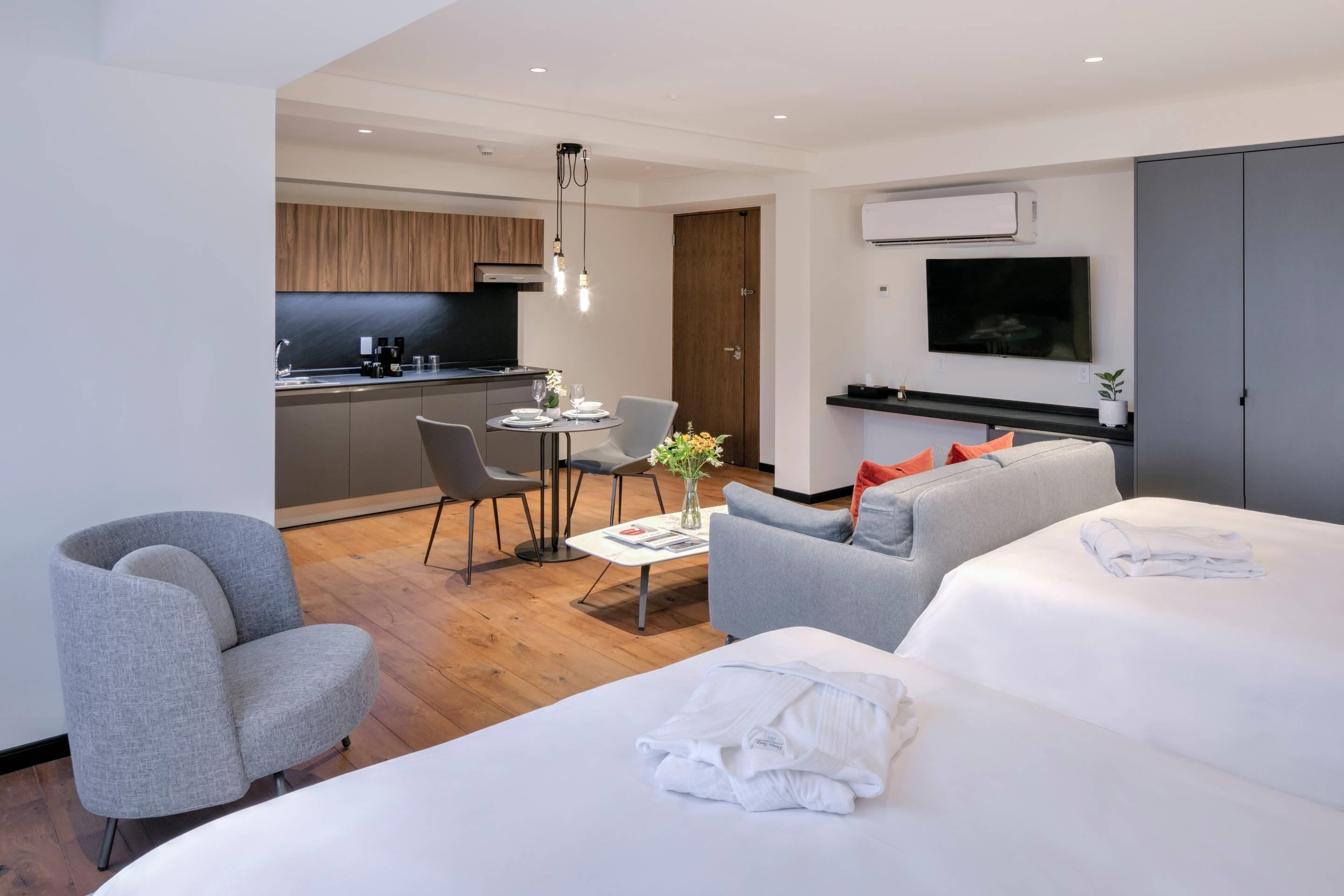 A modern hotel room with a large bed, a grey sofa, a dining area with a table and chairs, a kitchen in the background, and a TV mounted on the wall.