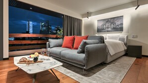 A modern living room with a grey sofa, red and white pillows, a marble coffee table with snacks and drinks, a grey rug, and a cityscape view through a large window. - Amberes 64, an Ascend Collection Hotel (Mexico City)