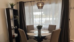Uma sala de jantar com uma mesa redonda de vidro, quatro cadeiras, um vaso com flores e uma luminária pendente.