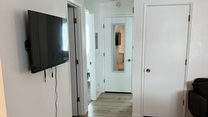 Um corredor com televisão de tela plana na parede, uma porta com espelho e um sofá de cor escura.