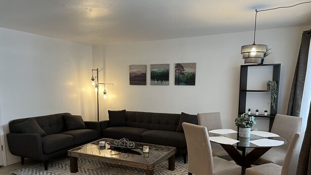Uma sala de estar moderna com um sofá escuro, uma mesa de centro de vidro e uma mesa de jantar com cadeiras. Há luminárias fixadas na parede e quadros emoldurados.