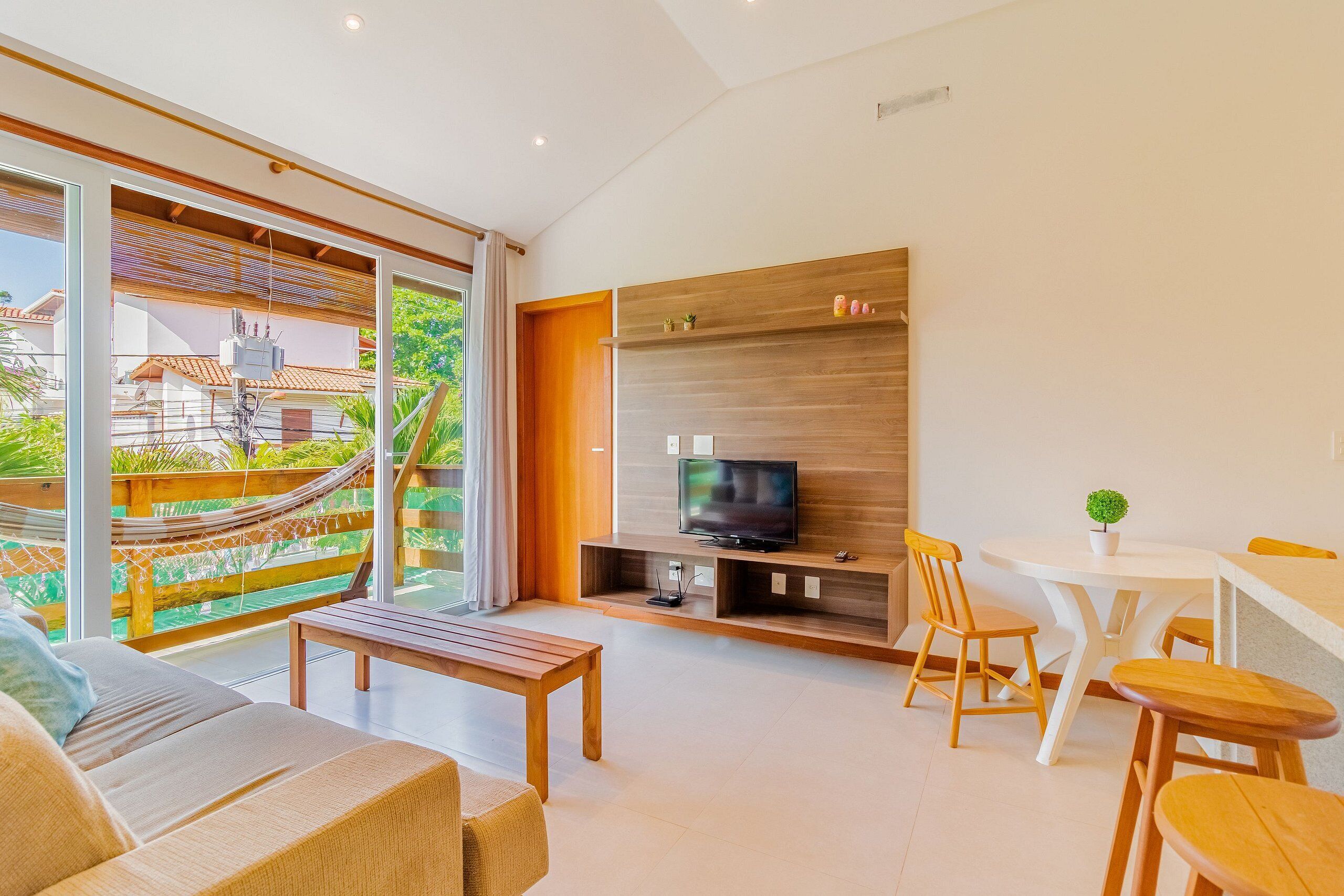 Una sala de estar moderna con un sofá, un banco de madera, un televisor sobre una estantería de madera y una zona de comedor con sillas y una mesa.