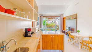 A modern kitchen with a central island, a dining area with wooden chairs, and a view of a garden through large windows. - PF49 Apto Aconchegante a 200m do Mar (Mata de São João)