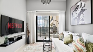 A modern living room with a large flat-screen TV, a sofa with colorful pillows, a coffee table, and a view of a cityscape through a large window. - Heavenly New 1 BD in Business Bay (Dubai)