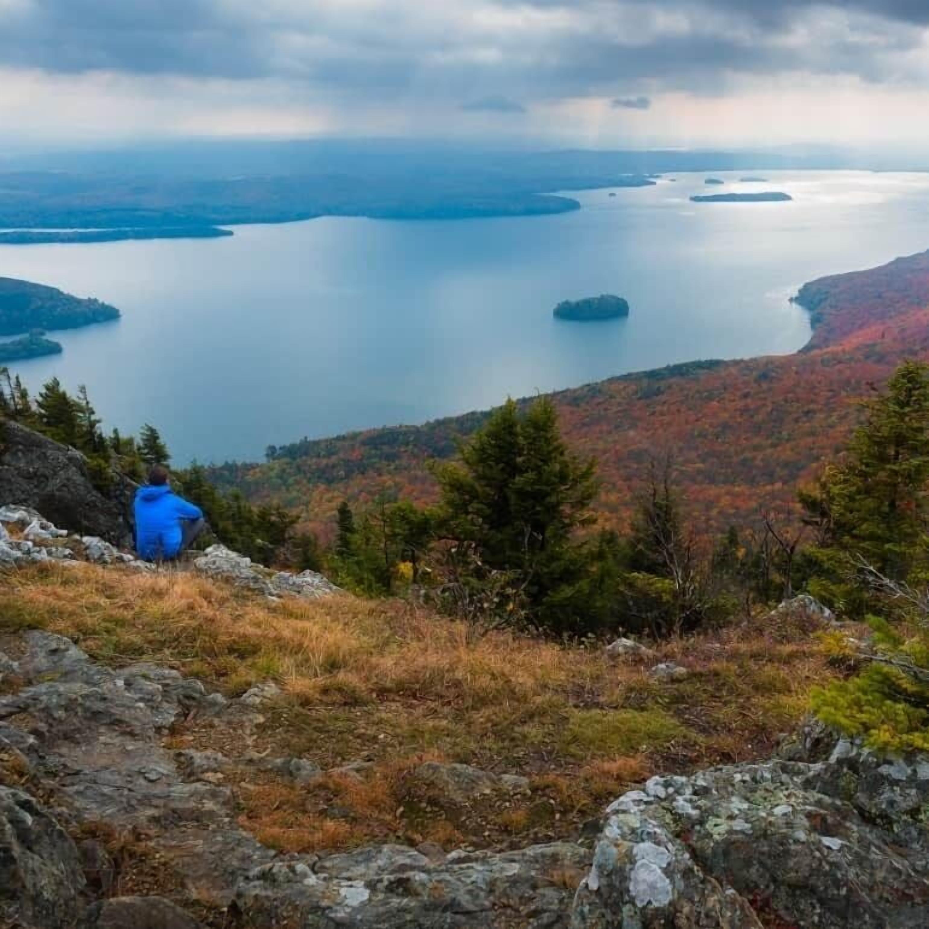 Eine Person in einer blauen Jacke sitzt auf einer felsigen Klippe mit Blick auf einen See mit mehreren Inseln, umgeben von herbstlich gefärbten Bäumen.
