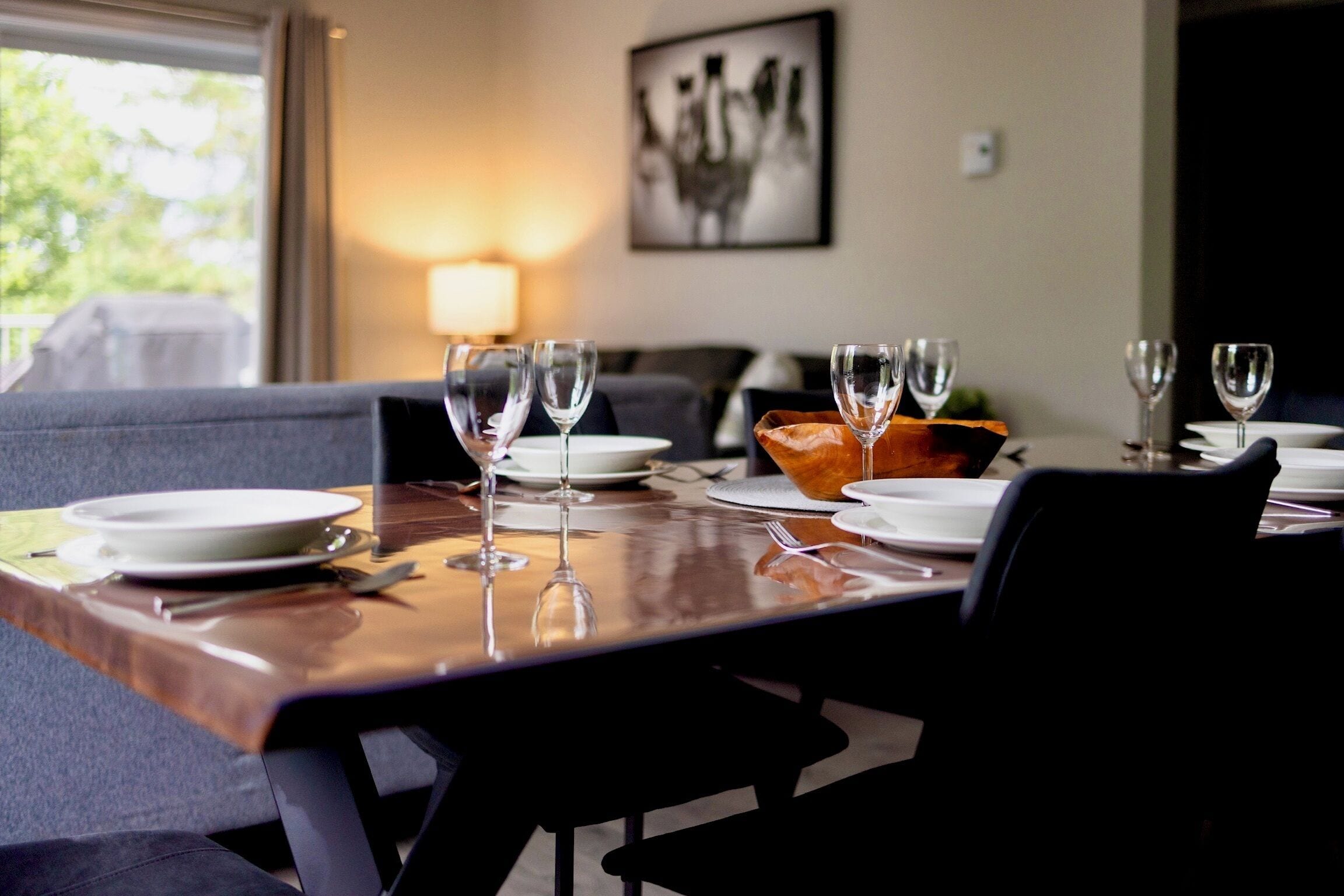 A dining table set with plates, cutlery, and wine glasses, with a bowl of bread and a lamp in the background.