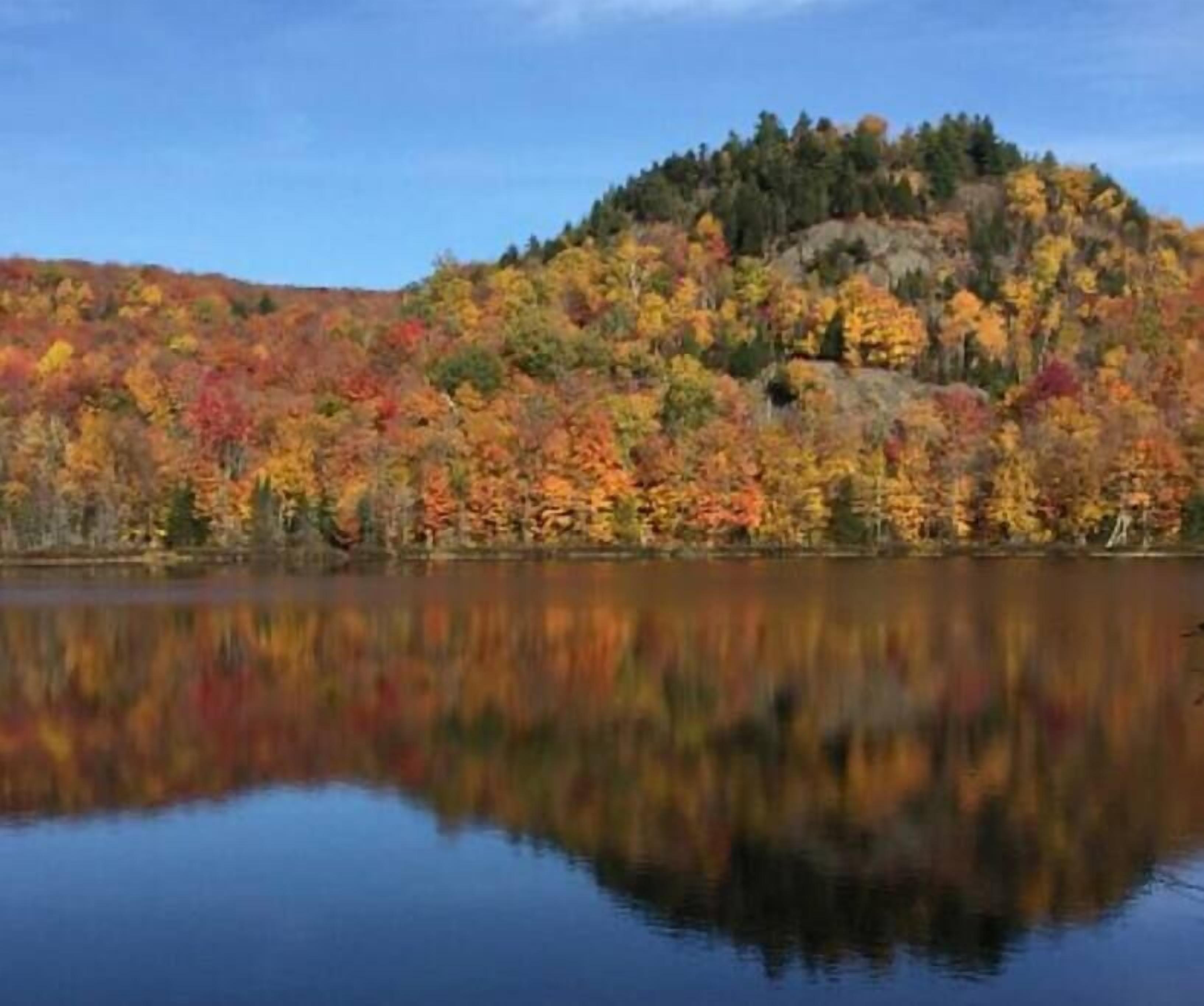 A calm lake reflecting a forested hill with autumn foliage.