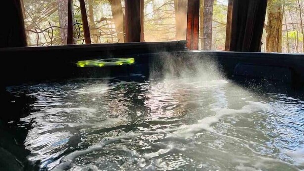 Una fuente termal con vapor saliendo del agua, rodeada de vigas de madera y vistas a árboles a través de una ventana.
