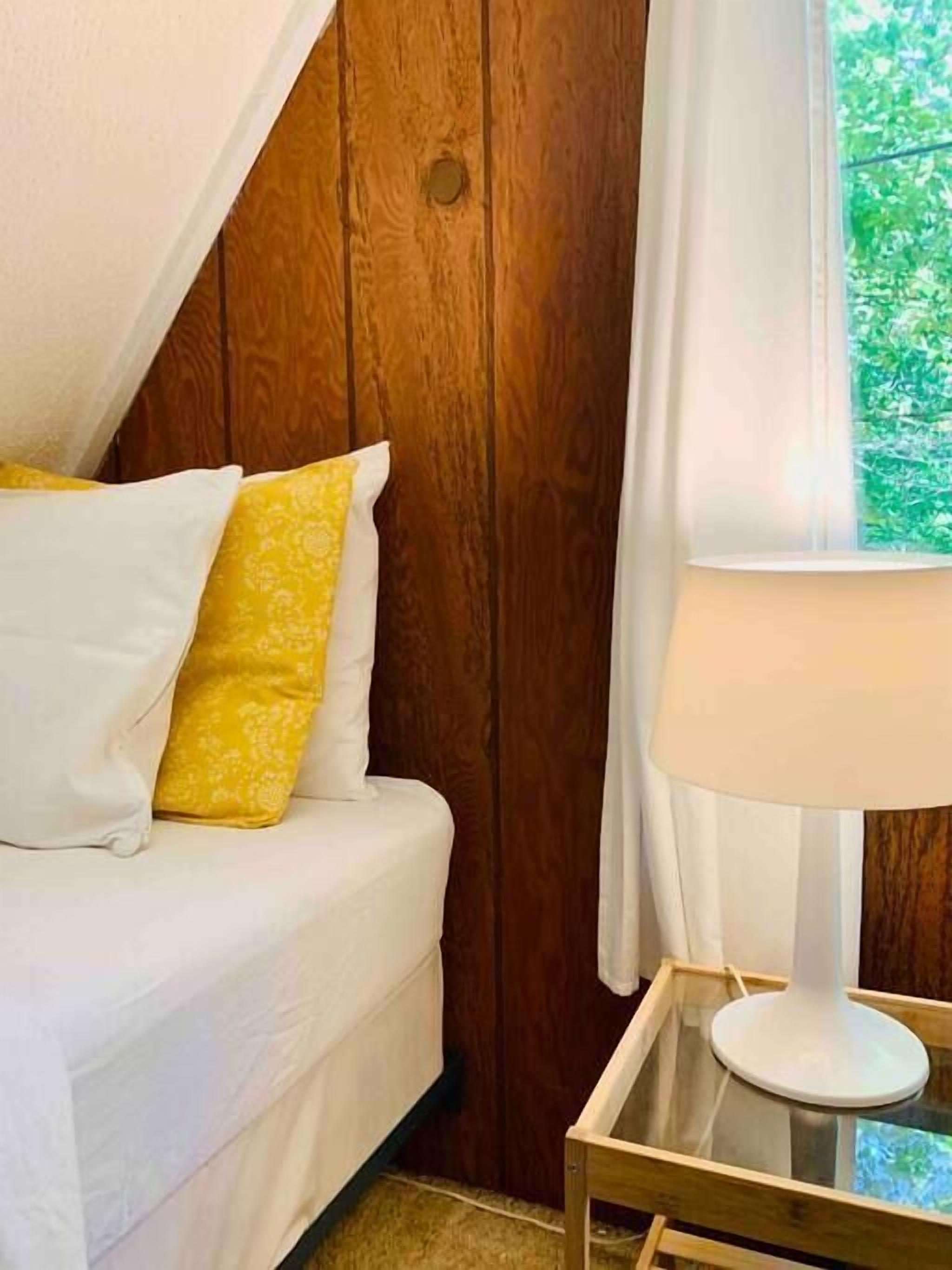 Un dormitorio con cabecera de madera, cama blanca, almohada amarilla, lámpara blanca sobre una mesa de vidrio y ventana con vistas a la vegetación.