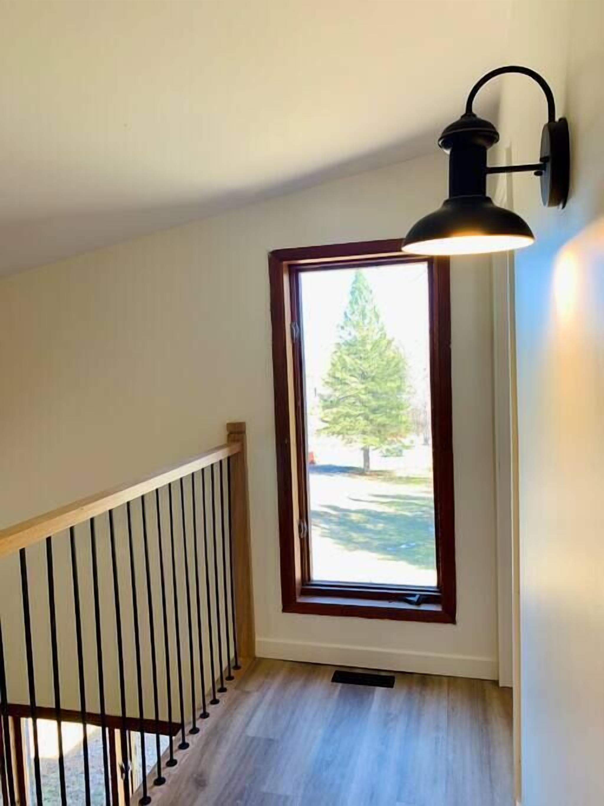 Un escalier avec une rampe en bois et un luminaire noir fixé au mur, situé au-dessus d’une fenêtre donnant sur un arbre.