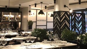 Un interior de restaurante con mesas dispuestas para comer, una zona de bar con botellas, y un letrero que dice "ALESSA".