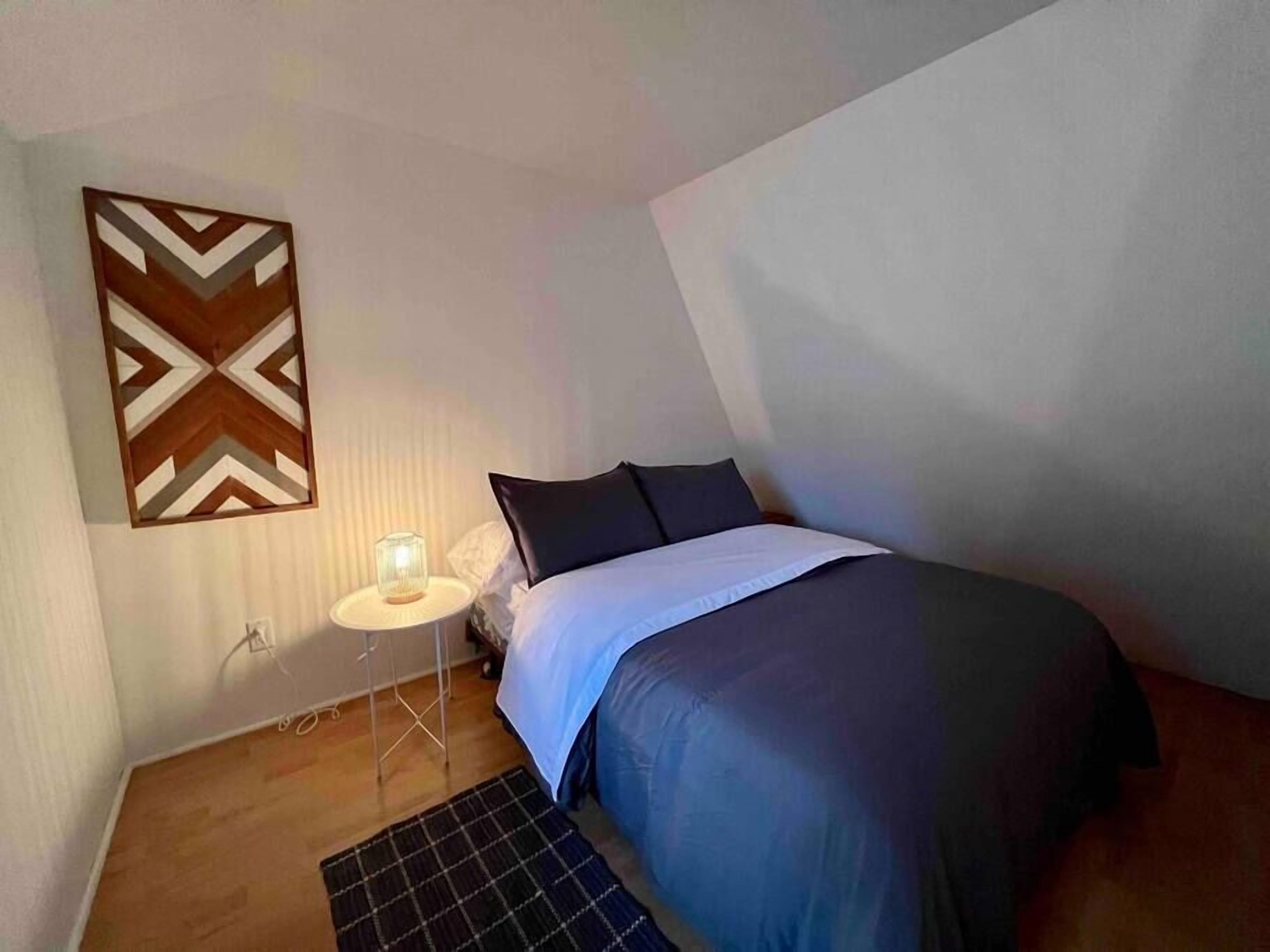 Un dormitori amb un llit, una tauleta de nit amb una làmpada, una decoració mural amb motius i un sòl de fusta.