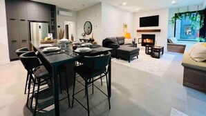 Ein modernes Wohnzimmer mit Kamin, eine Essecke mit schwarzem Tisch und Stühlen sowie eine Küche mit integriertem Kühlschrank.