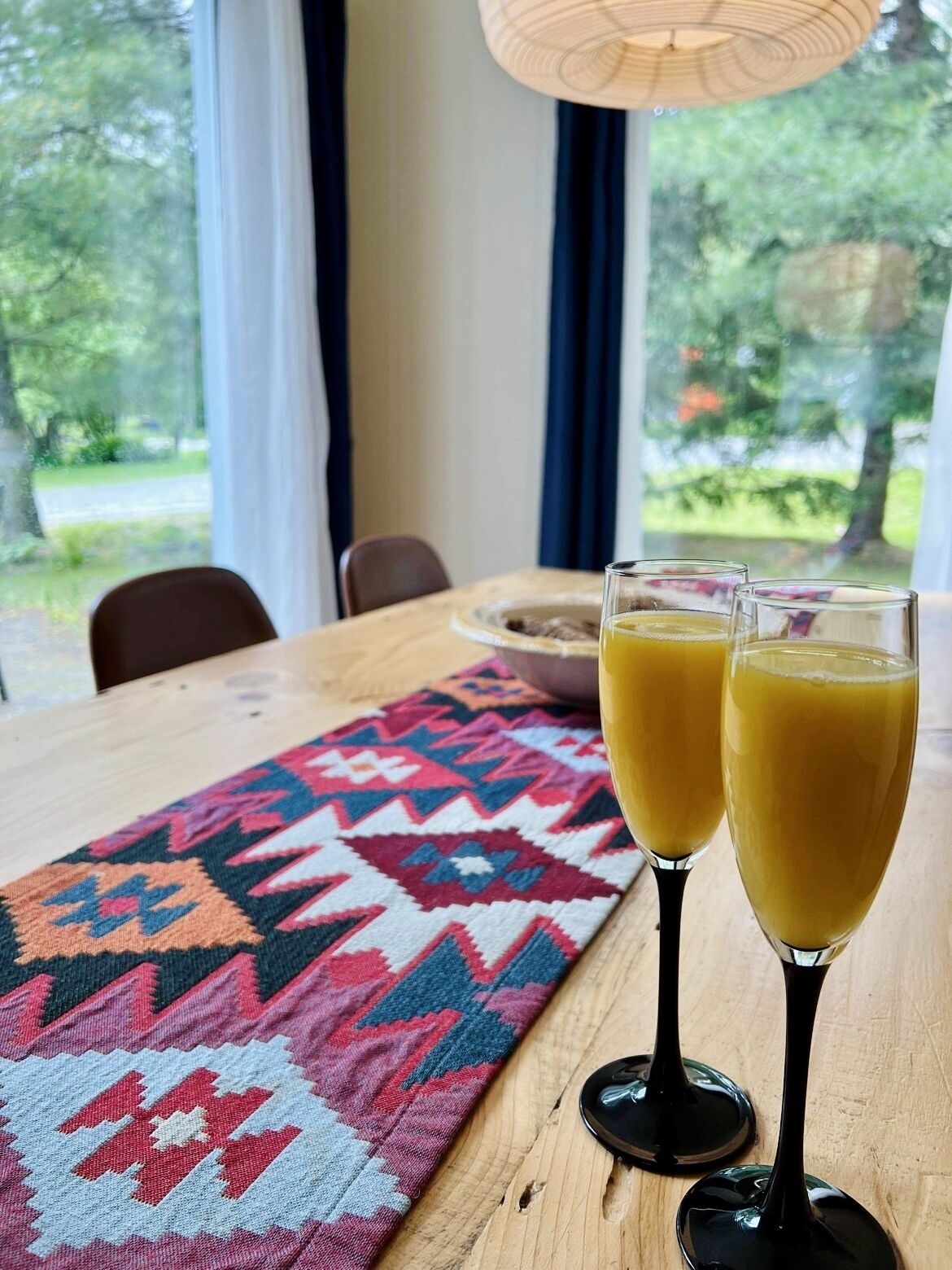 Uma mesa de jantar com dois copos de sumo de laranja, um tapete estampado e vista para o verde através de uma janela.