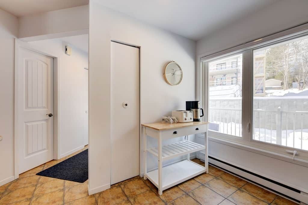 A hallway with a white door, a small shelf with a coffee maker, and a window with a view of a balcony.