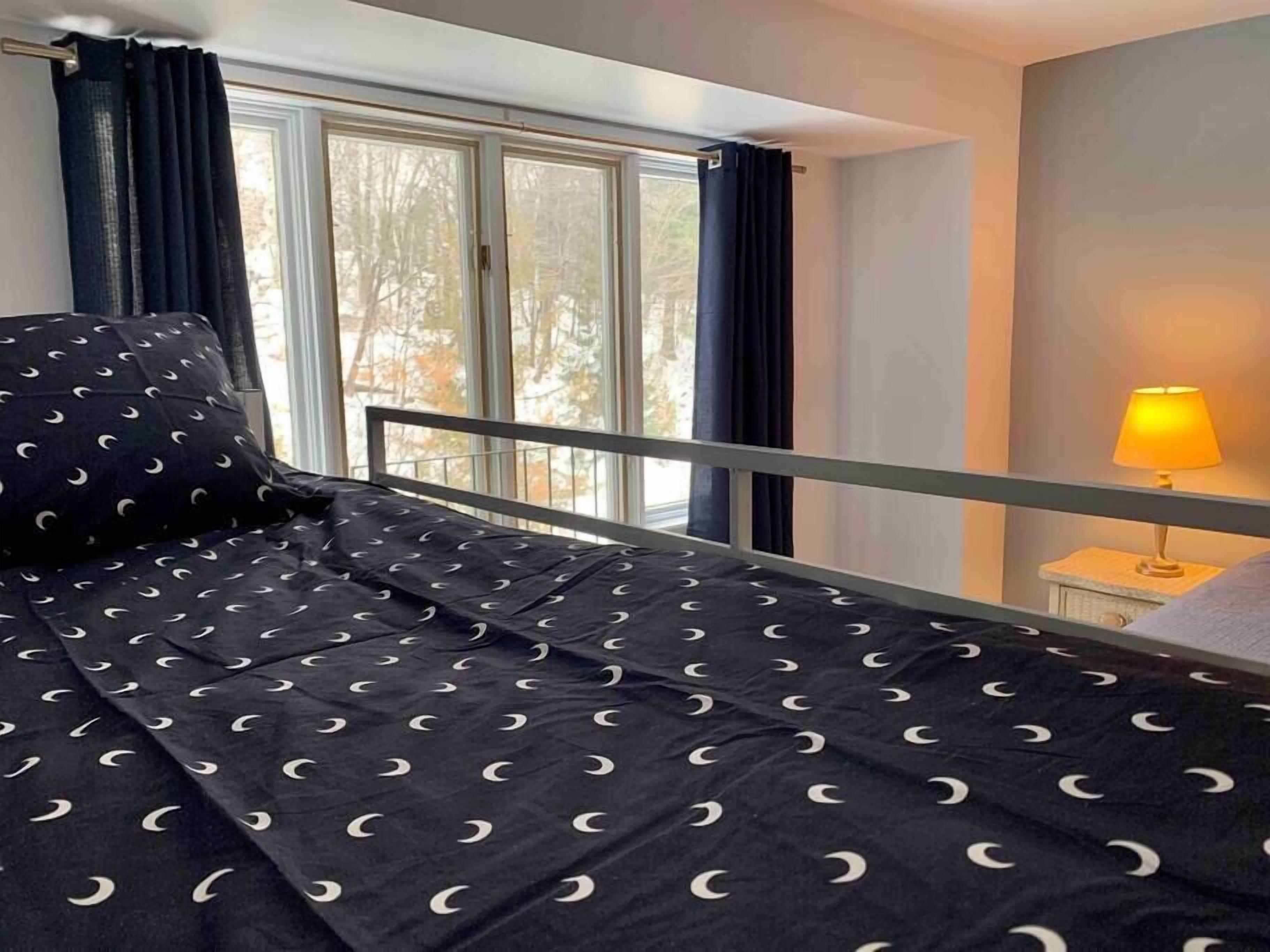 Ein Schlafzimmer mit Bett, einem Fenster mit Vorhängen, einem Nachttisch mit Lampe und Blick auf Bäume.